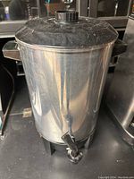 Stainless steel percolator with black plastic handles and lid, equipped with a spigot for coffee dispensing.