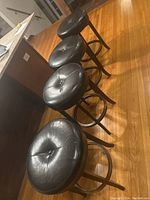Photo showing four black leather-look round bar stools with dark wood legs positioned in a row. Tops exhibit wear and some splitting.