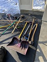Group of used garden and yard tools including shovels, rakes, axes, and a snow shovel placed on driveway outside with snow nearby.