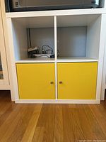 Front view of white IKEA Kallax cube storage unit with 2 yellow doors on the bottom row and 2 open cubes on top.