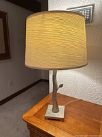 Lamp showing textured beige shade illuminated, mounted on a sculpted tree branch-like metallic base on a wooden table.