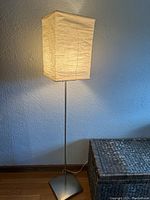 Lit floor lamp with square white paper shade and slim metal stand shown against a wall with wooden floor