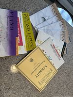 Piano tutorial books laid out on a surface showing titles such as 'Piano Adventures,' 'Pageants for Piano,' 'The Piano Course,' and 'Schmitt Preparatory Exercises.' The books are beginner-level piano learning materials.