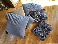 Top view of 7 pillows of various sizes and textures arranged on wooden floor including smooth and ruffled chenille pillows in grey and beige tones.