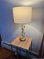 Full view of the table lamp on a wooden surface under power showing the illuminated white fabric shade and brass base with acrylic block details.