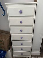 Front view of tall white wooden dresser with seven drawers and purple knob handles.
