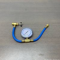 Photo of blue refrigerant charging hose with brass can tap valve and pressure gauge, laid on gray surface.