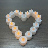24 white plastic flameless LED tea lights arranged in a heart shape on a dark wooden surface, some lights turned on emitting a warm yellow glow.