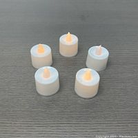 Five white plastic flameless LED tea lights showing synthetic flame tip and soft flickering glow on gray textured surface.