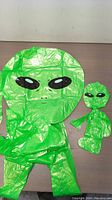 Two inflated alien figures, one large and one small, showing the bright green color, black eyes, and overall shape.