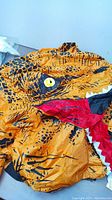 Close-up of the dinosaur face section showing yellow eye, red mouth, white teeth and black spots on orange inflatable fabric.