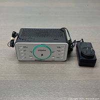 Front view of white and black Itoma alarm clock showing circular wireless charging pad with glowing light and multiple function buttons on the front panel, with power adapter to the side.