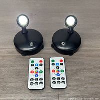 Two black RGB cordless spotlight units emitting bright white light, each paired with its remote control placed below the units on a flat surface. The spotlight heads are adjustable and pointed upward.