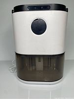 Front view of white compact dehumidifier showing black top panel and circular window. Bottom water tank dark smoke colored and transparent for water visibility.