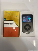 MP3 player inside its box next to colorful packaging with grid pattern and 'Music Player' label.
