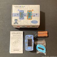 Photo of the Volueth Fingertip Pulse Oximeter in blue rubber protective case alongside user manual, two AAA batteries, blue lanyard, and product box showing front design with device illustrations and branding.