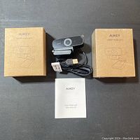 Two boxed Aukey 1080p webcams, one unboxed webcam showing USB cable and clip, and user manual on black surface