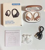 The earbuds inside the charging case, box showing 'Sweat Proof Comfortable to Wear' with super long battery life claim, user manual, USB charging cable, and extra ear tips in a plastic bag.