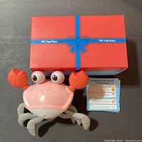 Wireless Crawling Crab Toy 2.0 with pink body, red claws and eyes, accompanied by instruction manual and red box with blue ribbon.