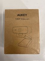 Front view of sealed brown box labeled Aukey 1080P Webcam with line art of the webcam on front.