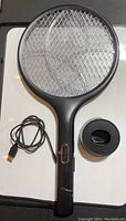Full view of the black bug zapper racket alongside a USB charging cable and a circular charging base, all placed on a white surface.