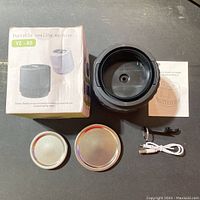 Photo showing black electric vacuum sealer, two metal lids, manual, USB cable, and small tool on a dark table with packaging box in background.