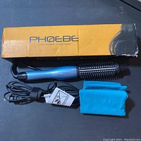Phoebe hair straightener brush in blue with power cord next to original yellow and black box and two turquoise pouches on flat surface.