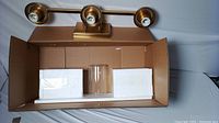 3-light brushed brass vanity wall fixture with bulb sockets and clear glass shade visible, in open box with foam packaging