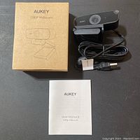 Photo of the Aukey 1080P webcam with attached USB cable, adjustable clip stand, user manual, and the original brown cardboard box with product illustration and branding on a flat surface.