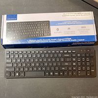 Front view of the Insignia Bluetooth Full-Size Keyboard alongside its retail packaging box showing key features such as Bluetooth compatibility, 104 keys, scissor switch technology, and multi-OS support.