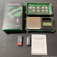 Box, scale, batteries, calibration weight, and instruction manual laid out on black surface.