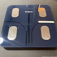 Top view of BobBros smart scale showing dark glass platform and four metallic sensor electrodes.