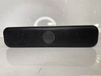 Front view of the black USB computer soundbar speaker with a central circular control button embossed with an X symbol, placed on a reflective white surface.