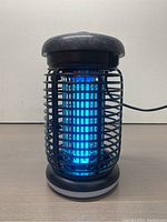 Front view of black cylindrical bug zapper illuminated with blue UV light.