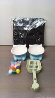 Photo showing all items included: two blue ceramic cat bowls, green plastic cat litter scoop, black textured cat litter mat in packaging, and pack of multicolored pom pom toys.