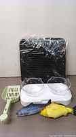 Adjustable double pet bowls with clear cat-shaped dishes on white base, green litter scoop, black textured litter mat, and two fish-shaped toys.