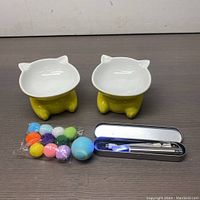 Photo showing two yellow ceramic cat-shaped bowls with white interiors, packaged multi-colored pom pom balls, blue-white spherical cat toy ball, and metal case containing silver laser pointer and USB cable.