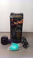 Photo showing the Conair Curl Secret automatic hair curler, its original box, power cord, and a turquoise silicone scalp massager in front on a wooden surface.