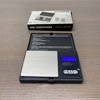 Digital professional mini scale open showing stainless steel weighing platform and blue backlit digital screen, with buttons and instruction text in lid. Original box behind scale.
