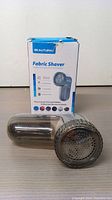 Fabric shaver placed in front of its original box showing front and side views and lint compartment.