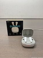 White Gummi Itoma wireless earbuds seated inside open charging case, red LED indicator light visible on case front, original product box displayed behind on table.