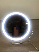LED vanity mirror powered on with cool white LED lights illuminated around the perimeter. The mirror has a clear reflection and stands on a white foldable stand with a visible charging cable.