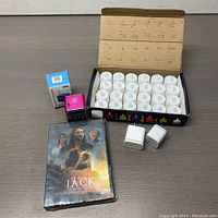 Photo showing the open box of 24 flameless LED tea light candles, the sealed DVD titled 'When Jack Came Back', a small pink Bluetooth speaker, and two white plug adapters on a wooden surface.