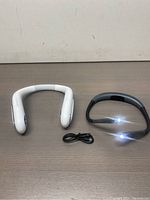 White U-shaped portable neck fan and black LED neck light with USB charging cable on table.