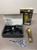 Gold professional hair clipper with box, charger, multiple clipper guide combs, scissors in plastic, and manual laid out on wooden surface.