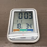 Front view of Geevon digital bathroom clock showing timer at 3 seconds, 18.0°C temperature, 34% humidity, and moisture level indicator bar. Buttons visible at bottom for timer control and clear.