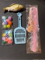 Wide shot showing fish toy, pom-poms, litter scoop, and pink feather wand on black surface