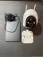 Photo showing white solar security camera with dual antennas, black front sensor panel, power adapter, and mounting bracket on a table.