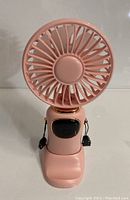 Front view of the pink handheld fan showing circular fan grille, power button, and foldable base.