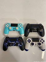 Lot of four PS4 style video game controllers in various colors and designs on white surface.